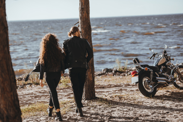 back view of couple holding hands and walking on seashore, cruiser motorcycle standing near - Zdjęcie, obraz
