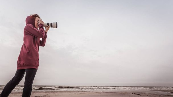 Woman having leisure time walking on coastline during autumnal weather taking pictures using camera - Фото, изображение