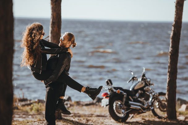 happy man holding his girlfriend on arms on seashore with classical motorcycle near - Photo, Image