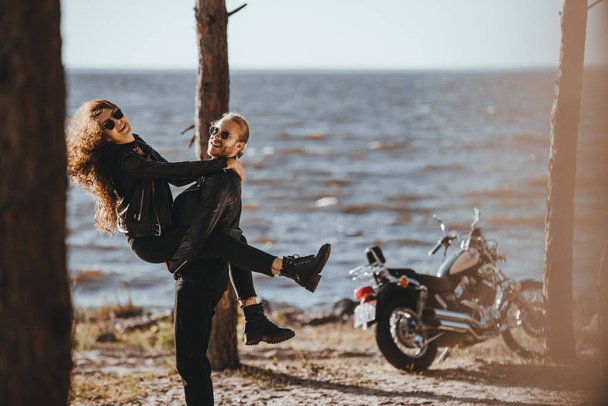 boyfriend having fun and holding his girlfriend on arms on seashore with motorcycle near - Photo, Image