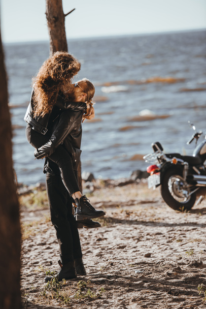 мужчина держит свою девушку на руках на берегу моря с мотоциклом рядом
 - Фото, изображение