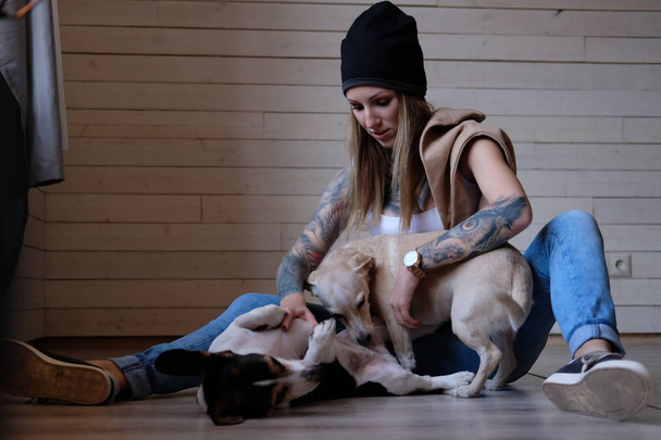 Portret van een hipster tattoed meisje speelt met haar kleine honden zittend op een houten vloer. - Foto, afbeelding