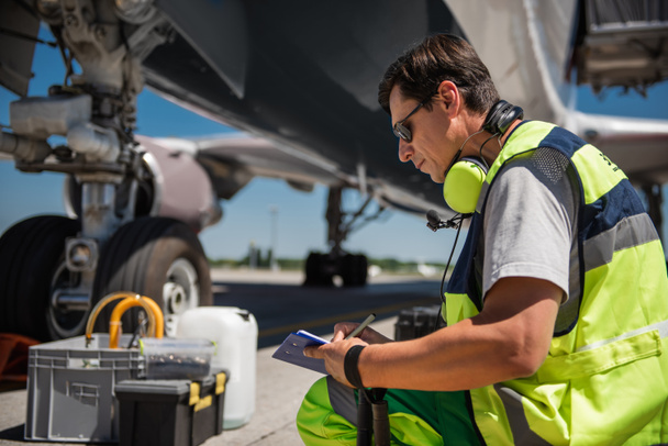 Pracownik lotniska w kucki w pobliżu samolotów i pisanie w schowku - Zdjęcie, obraz