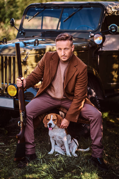 Hunter en ropa elegante sostiene una escopeta y se sienta junto con su perro beagle mientras se apoya contra un coche militar retro en el bosque
. - Foto, imagen