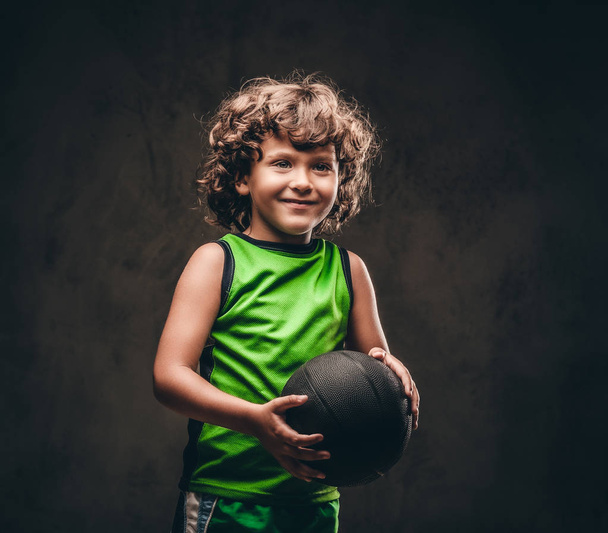 Little basketball player in sportswear holding ball in a studio. Isolated on a dark textured background. - Photo, Image