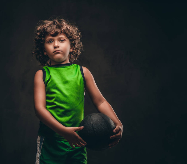 Portrait of a cute little basketball player in sportswear holding the ball in a studio. Isolated on a dark textured background. - Foto, immagini