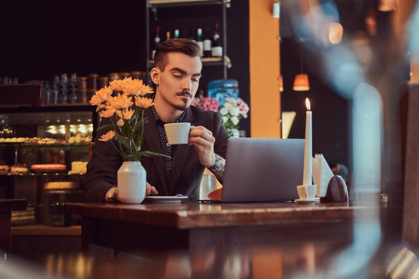 Hombre freelancer guapo con barba elegante y cabello vestido con un traje negro que trabaja en el ordenador portátil mientras está sentado en un café
. - Foto, imagen