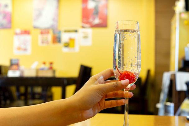 Femme main tenant un verre de champagne avec cerise au restaurant avec mur jaune
. - Photo, image