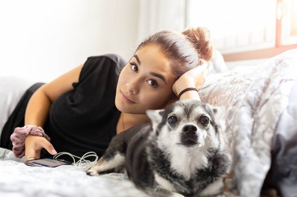 Mezcla de raza adolescente acostada en la cama con su perro chihuahua mirando a la cámara
. - Foto, imagen