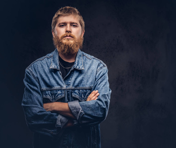 Portret van een knappe hipster man gekleed in jeans jasje poseren met gekruiste armen. Geïsoleerd op een donkere achtergrond. - Foto, afbeelding