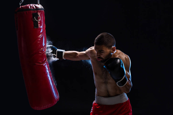 Bearded male boxer training with punching bag on black background. Young boxer training on punching bag. Male boxer as exercise for the big fight. Boxer hits punching bag. - Foto, Imagem
