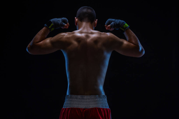 Rear view of strong young male boxer. Fitness male model wearing boxing gloves standing on black background. - Photo, Image