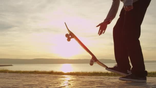 SLOW MOTION CLOSE UP Молодий скейтбордист розважається, граючи зі скейтбордом, кидаючи його в повітря і стрибаючи на ньому на схід сонця. Невпізнаваний ковзаняр робить трюки на бетонній береговій лінії
 - Кадри, відео