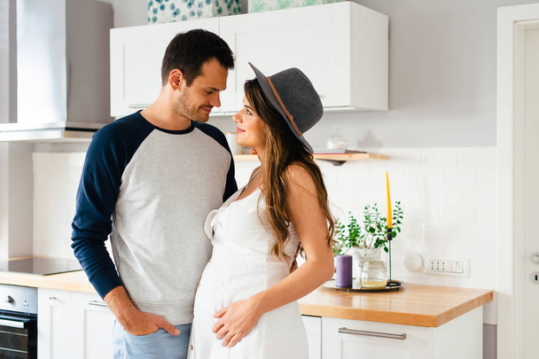 schönes junges modernes Paar erwartet ein Baby und schaut einander in der heimischen Küche mit Liebe an. - Foto, Bild
