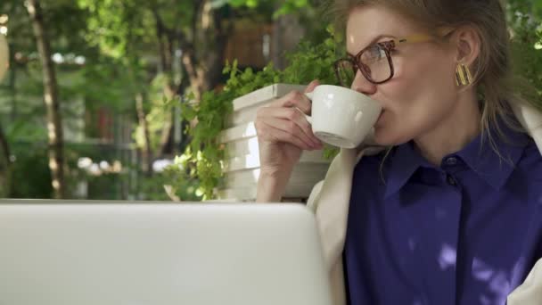 Attractive young hipster girl wearing glasses and a dark blue shirt is sitting in an outdoors cafe drinking hot beverage. A laptop. Handheld real time establishing shot - Séquence, vidéo