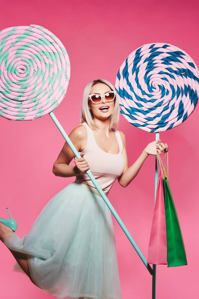 Attractive young woman wearing sunglasses holding huge sweet lollipops and shopping bags posing at pink background, shopping concept. - Photo, Image