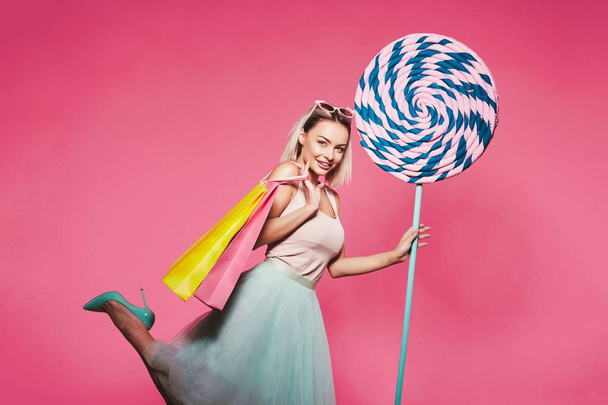счастливая блондинка держит огромный сладкий леденец и сумки для покупок позировать на розовом фоне, концепция покупок
. - Фото, изображение