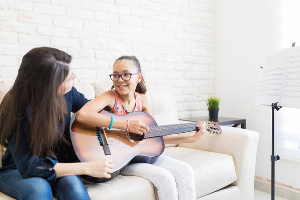 Female teacher teaching girl to play guitar while sitting on sofa at home - Photo, Image