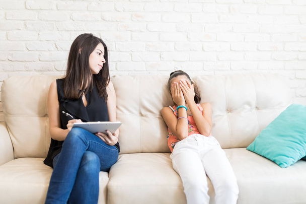 Psicólogo feminino olhando para menina triste cobrindo rosto com as mãos enquanto sentado no sofá em casa
 - Foto, Imagem