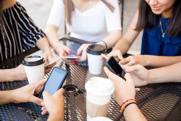 Ryhmä ystäviä istuu ulkona kahvilassa ja käyttää puhelimia, kuppeja kahvia pöydällä
 - Valokuva, kuva