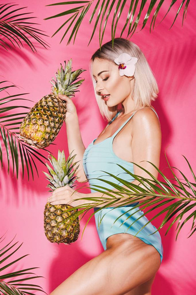 Attractive young girl with blonde hair wearing blue swimming suit standing with pineapples on pink background with palm leaves - Фото, изображение