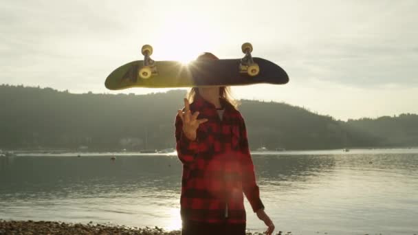 Zeitlupe, Nahaufnahme: Cooler lächelnder Skateboarder, der Spaß daran hat, mit seinem Skateboard zu spielen, es in die Luft zu werfen und es über die Sonne in die Kamera zu schieben. Verspielter Skater spielt mit Skateboard - Filmmaterial, Video