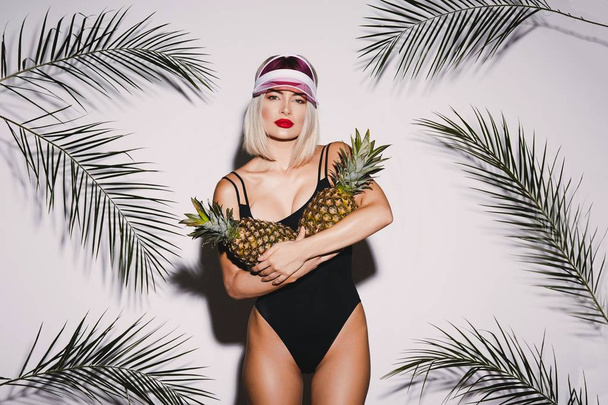 Attractive young blonde girl wearing swimming suit standing with two pineapples at white studio background with palm leaves - Foto, Bild