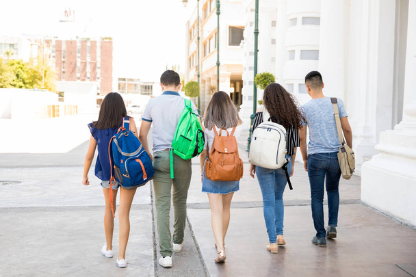 Vista trasera del grupo de cinco adolescentes y niñas caminando por la calle de la ciudad con la bolsa de las escuelas
 - Foto, imagen