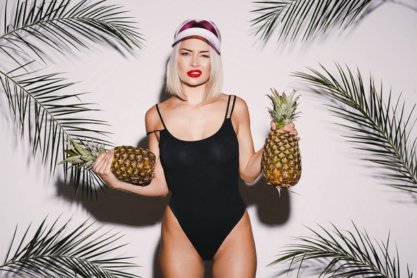 Cute girl with blonde hair wearing black swimming suit posing with pineapples - Photo, image