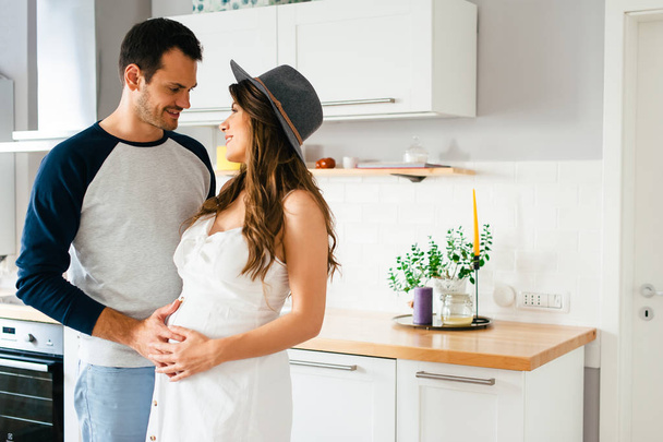 Krásná mladá moderní dvojice čeká dítě, s láskou při pohledu na sebe ve své domácí kuchyni, zatímco muž objímá svou těhotnou manželku. - Fotografie, Obrázek
