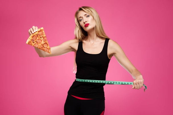 Mladá krásná smutná dívka na růžovém pozadí má pizzu v jejích rukou a měří v pase. Pojem nezdravá strava - Fotografie, Obrázek