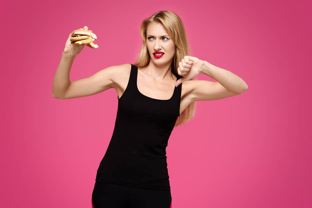 Jovem bela menina esbelta em um fundo rosa segura um hambúrguer em suas mãos e mostra o polegar para baixo. Conceito de junk food
 - Foto, Imagem