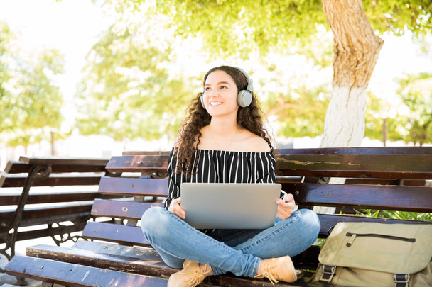 Портрет милой латинской девочки-подростка, сидящей на скамейке в парке с ноутбуком, смотрящей в камеру и улыбающейся
 - Фото, изображение