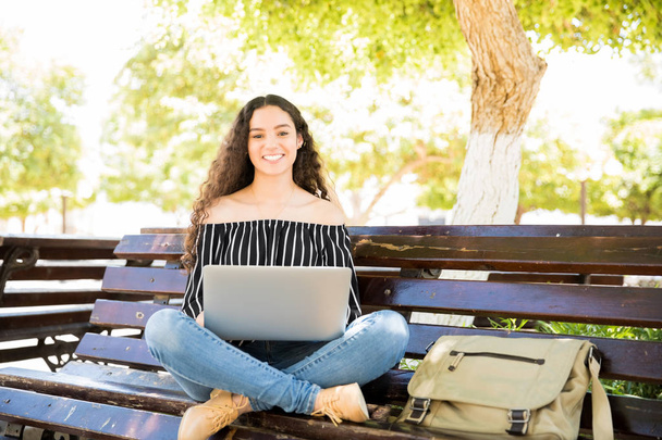 Portrait of cute latin teenage girl sitting on park bench with a laptop computer looking at camera and smiling - Photo, Image