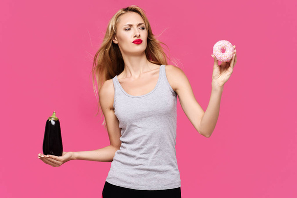Young beautiful slender girl with a pink background with a beautiful smile holding and looking to eggplant and donat.  Concept of un healthy fat junk and healthy food - Photo, image