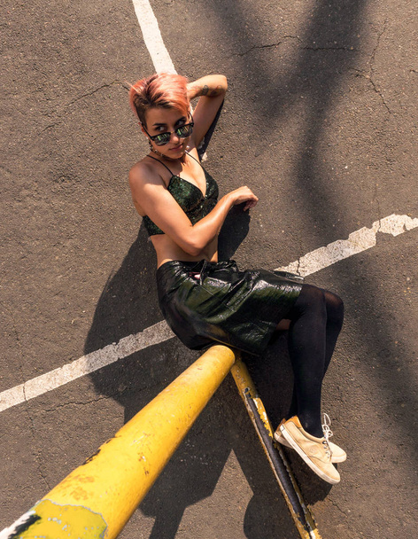 Foto in basso di Street punk girl con capelli rosa tinti Si siede su Iron Tube su sfondo asfalto. Donna con piercing passeggiare da parete a campo da basket con graffiti dipinti su pareti
 - Foto, immagini