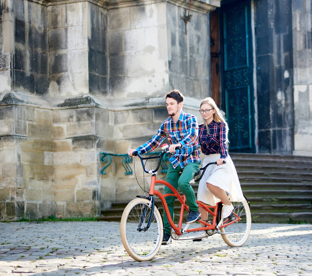 Jonge actieve gelukkig reiziger paar, knappe man met baard en mooie blonde vrouw in glazen fiets samen tandem langs verharde stoep op heldere zonnige Zomerochtend door oude gebouwen. - Foto, afbeelding