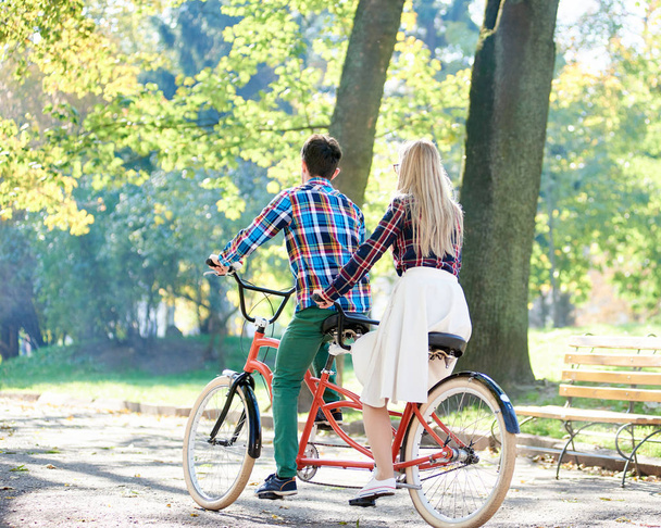 Back view of young active traveler couple, handsome man and attractive blond woman cycling together tandem double bike along crackled path in lit by bright sun beautiful park under tall trees. - Photo, Image