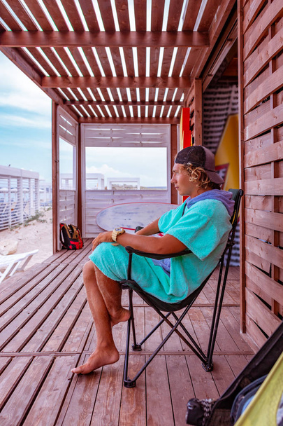 Ελκυστικός νεαρός σέρφερ φορώντας μπουρνούζι κάθεται στη βεράντα δίπλα στη θάλασσα, χαλαρωτικό και κοιτάζοντας την απόσταση στη θάλασσα - Φωτογραφία, εικόνα