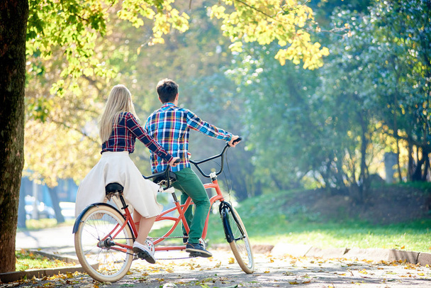 Back view of young traveler couple, handsome man and attractive blond woman cycling together tandem double bike along crackled path in lit by bright morning sun beautiful autumn park under trees. - Foto, imagen