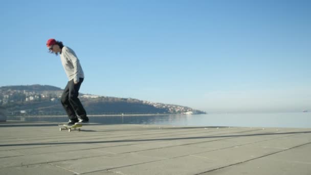 Zbliżenie w zwolnionym tempie: Młody pro Skater, skateboarding i skoki ollie flip podstęp przy promenadzie wzdłuż wybrzeża w słoneczne lato. Skater skoki kickflip trick z deskorolka, w pobliżu oceanu - Materiał filmowy, wideo