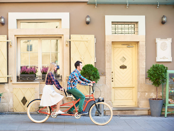 Side view of active tourist couple in casual clothing, bearded man and blond woman in glasses cycling tandem bike along empty paved sidewalk on bright sunny summer day by town residential houses. - Photo, Image