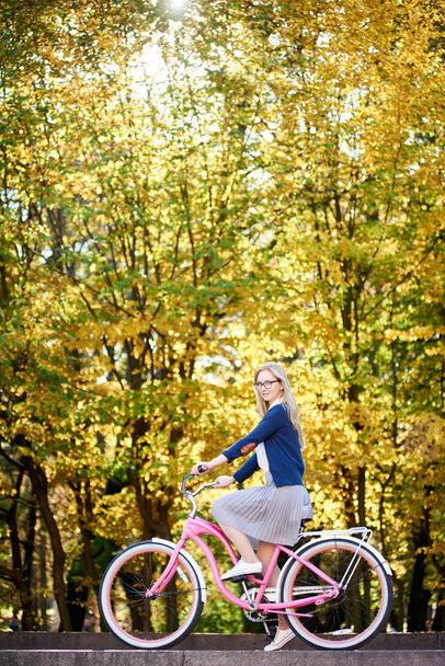 Vista lateral de la chica atractiva rubia activa de pelo largo en gafas, falda y blusa montando una moderna bicicleta rosa en iluminada por las escaleras del parque del sol de otoño sobre el fondo de los árboles bokeh dorados brillantes
. - Foto, imagen