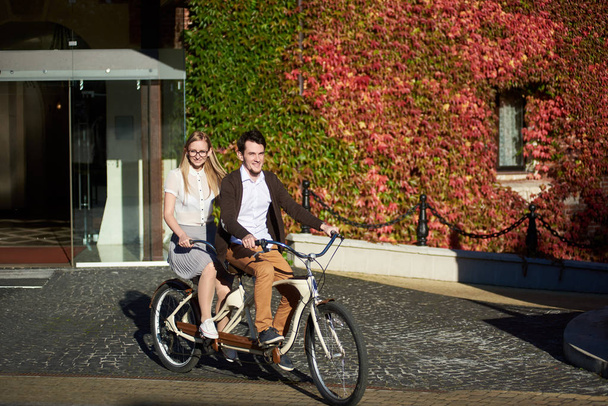 Young glimlachend toeristische paar, knappe man en mooie langharige blonde vrouw fietsen tandem fiets langs grijs straat op zonnige dag op achtergrond van bakstenen gebouw is begroeid met klimop rode bladeren. - Foto, afbeelding
