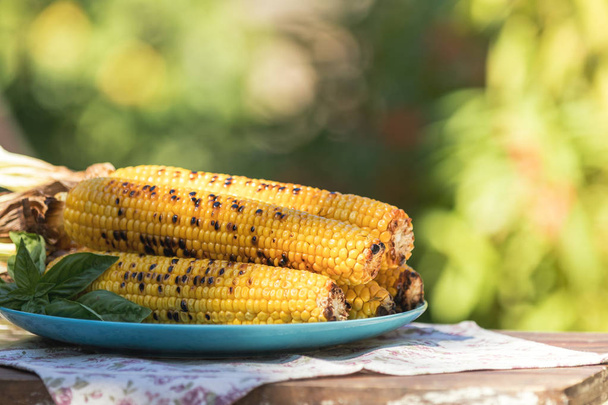 Corn baked in olive oil, with pepper, salt and basil on blue dish. Tasty grilled corn cobs on old table in the open air. Vegetarian, vegan menu - Photo, Image