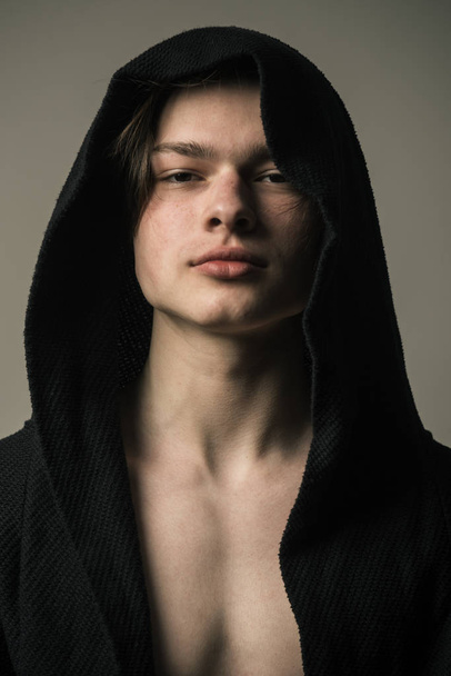 Guy on confident face in black hood, grey background. Man with smooth skin on face, neck and chest looking at camera. Macho in black bathrobe with hood, close up. Masculinity concept. - Foto, afbeelding