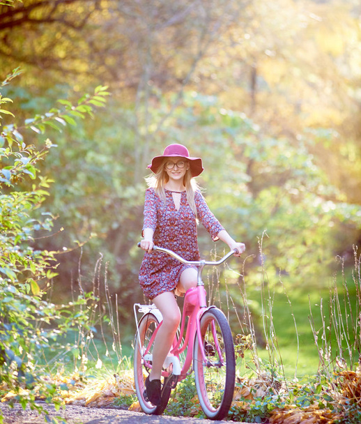 Slim happy smiling blond fashionable attractive girl in glasses, short dress and pink hat riding lady bicycle along paved park alley on beautiful green and golden trees lit by bright sun background. - Photo, Image