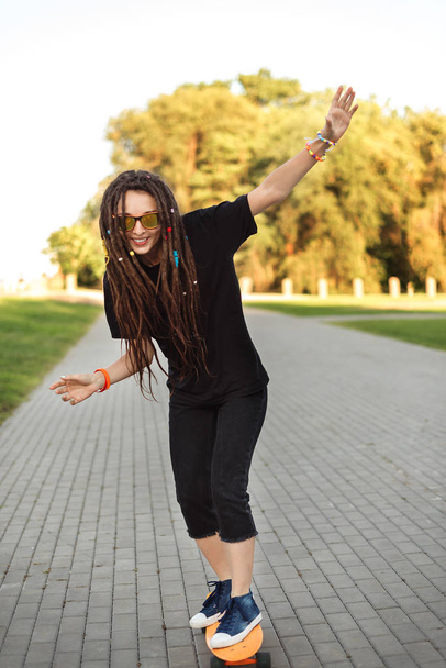 a girl in sunglasses is riding a longboard. young girl with dreadlocks. hippie on the skateboard keeps the balance - Foto, Bild