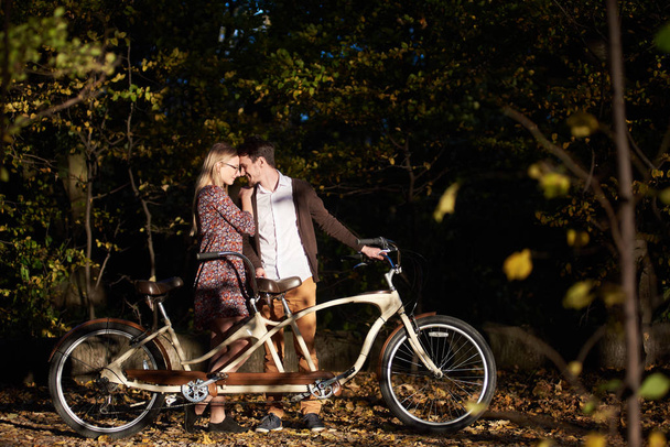 Young happy romantic couple, bearded man and attractive fashionable woman close together at tandem double bicycle outdoors in autumn park or forest on blurred dark trees dense foliage background. - Photo, Image