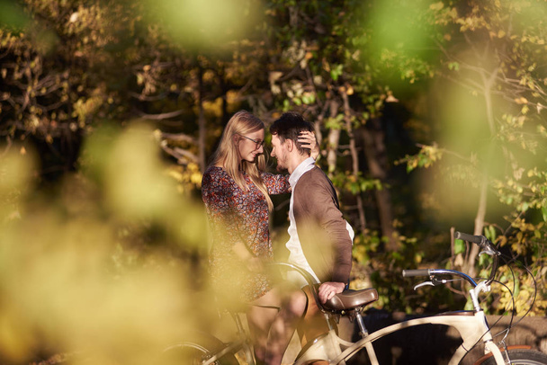 Casal romântico sorridente, homem barbudo e mulher atraente perto juntos na bicicleta dupla tandem ao ar livre no parque de outono escuro ou na floresta na clareira ensolarada no fundo bokeh verde dourado borrado
. - Foto, Imagem